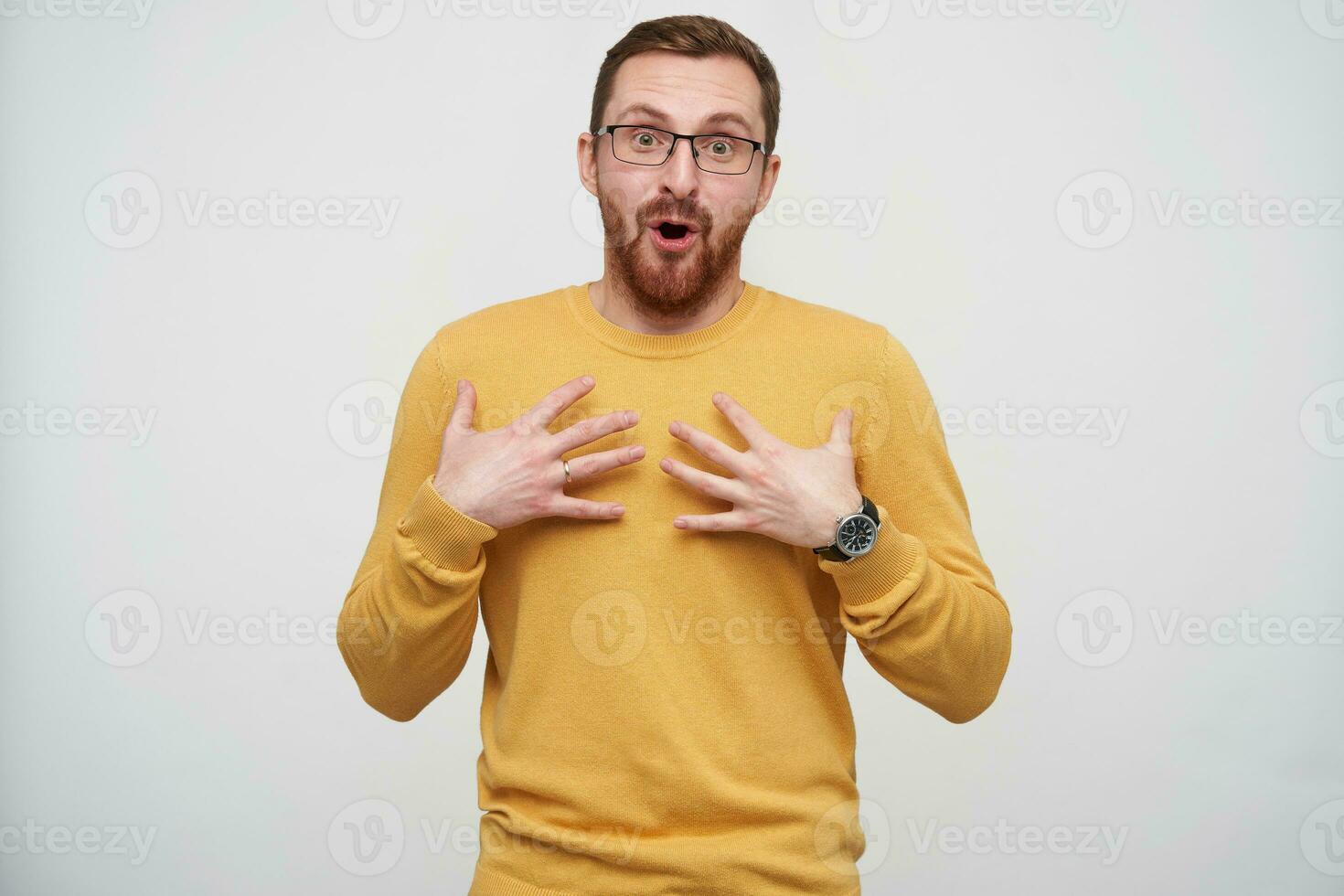 les yeux ouverts jolie Jeune barbu gars dans lunettes avec marron court cheveux en portant paumes sur le sien poitrine et à la recherche avec surprise à caméra, isolé plus de blanc Contexte photo