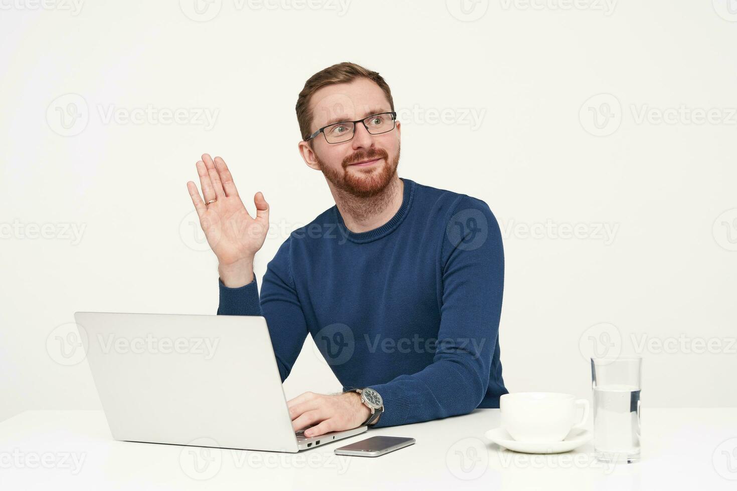 Beau Jeune non rasé blond homme habillé dans bleu chandail élevage paume dans Bonjour geste tandis que à la recherche de côté, travail avec le sien portable plus de blanc Contexte photo