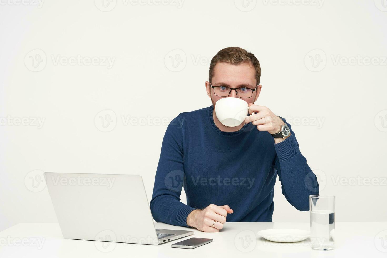 studio coup de Jeune barbu Masculin dans des lunettes ayant café Pause tandis que travail avec le sien portable et à la recherche à caméra, habillé dans bleu chandail tandis que séance plus de blanc Contexte photo