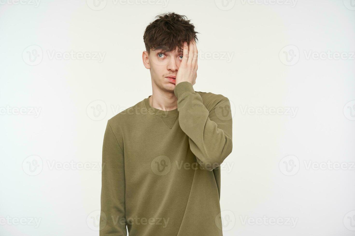 Jeune homme, fatigué à la recherche gars avec brunette cheveux, perçant et poil. portant kaki Couleur chandail. émouvant il est visage avec palmier, épuisé. en train de regarder à le caméra isolé plus de blanc Contexte photo