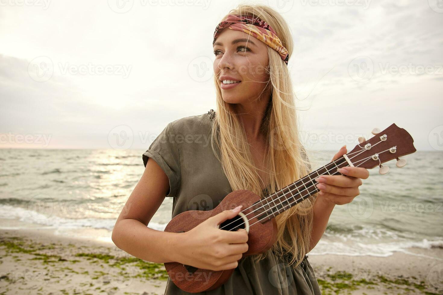 horizontal coup de Jeune attrayant à tête blanche femme dans Bandeau à la recherche volontier de côté avec large sourire et en gardant guitare dans sa mains tandis que posant plus de côte photo