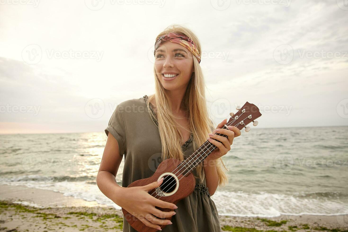 joyeux Jeune charmant longue aux cheveux blond femme avec Naturel maekup souriant largement tandis que à la recherche gaiement de côté, posant plus de plage Contexte avec ukulélé photo