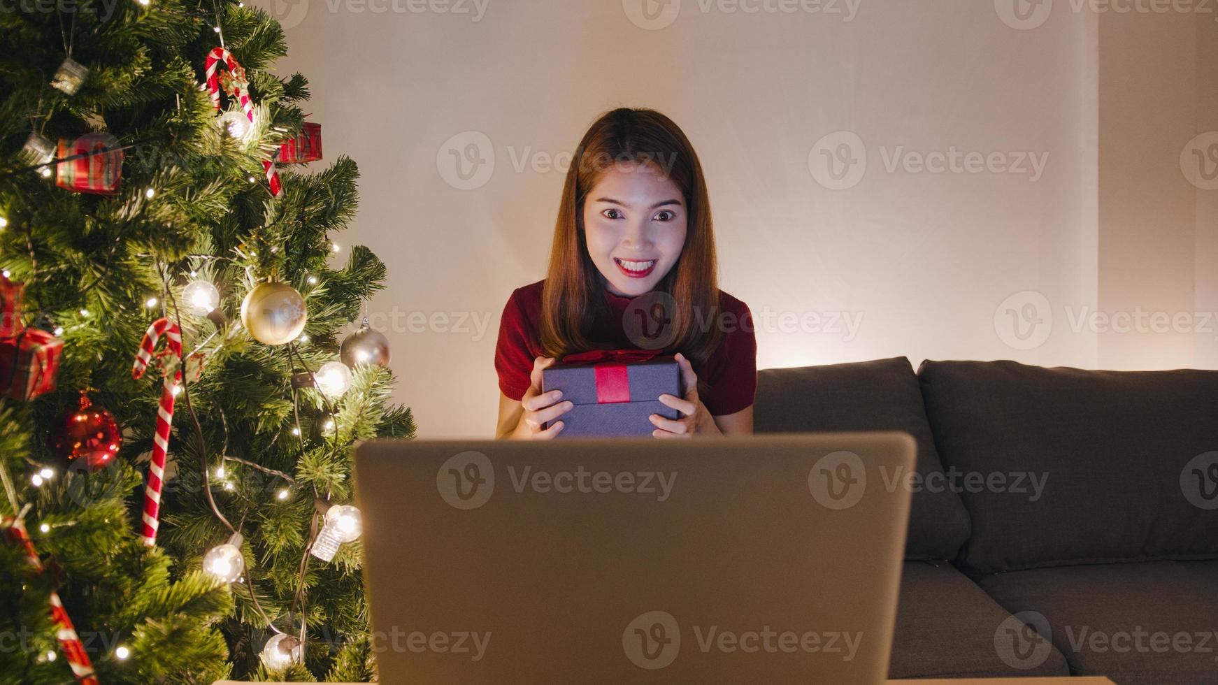 jeune femme asiatique utilisant un appel vidéo pour ordinateur portable parlant avec un couple avec une boîte cadeau de Noël, un arbre de noël décoré d'ornements dans le salon à la maison. nuit de noël et festival de vacances du nouvel an. photo
