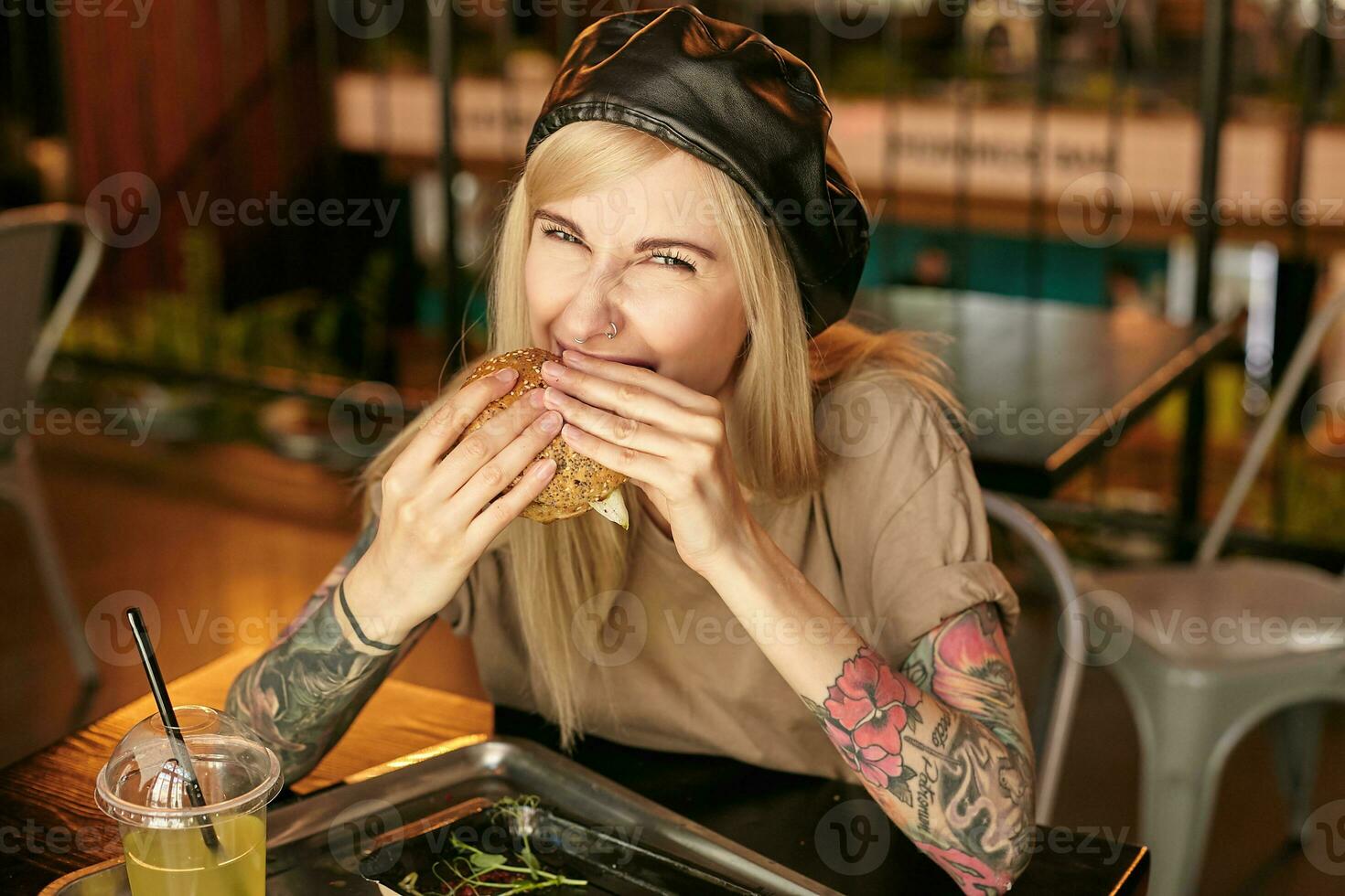 portrait de charmant de bonne humeur tatoué blond femelle avec décontractée coiffure portant beige T-shirt et cuir noir béret, séance à table dans ville café et en mangeant délicieux Burger photo