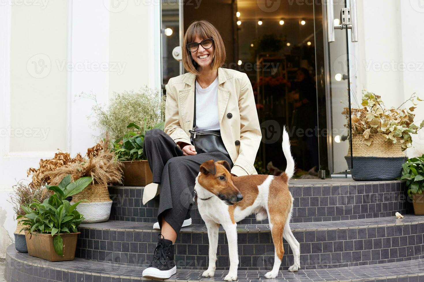 Jeune femme habillé mine de rien séance sur escaliers de fleurs magasin souriant à la recherche caméra, sa charmant animal de compagnie chien jack Russell terrier des stands proche, elle regards content, Extérieur photo, plus de rue Contexte photo