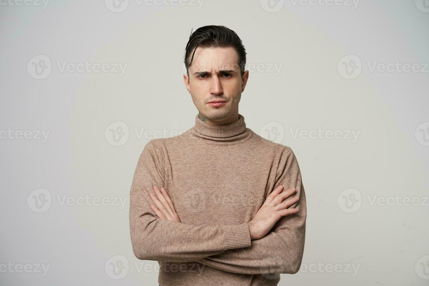 sévère Jeune foncé aux cheveux homme dans beige col roulé chandail permanent plus de blanc Contexte avec plié mains, à la recherche durement à caméra et renfrogné le sien les sourcils photo