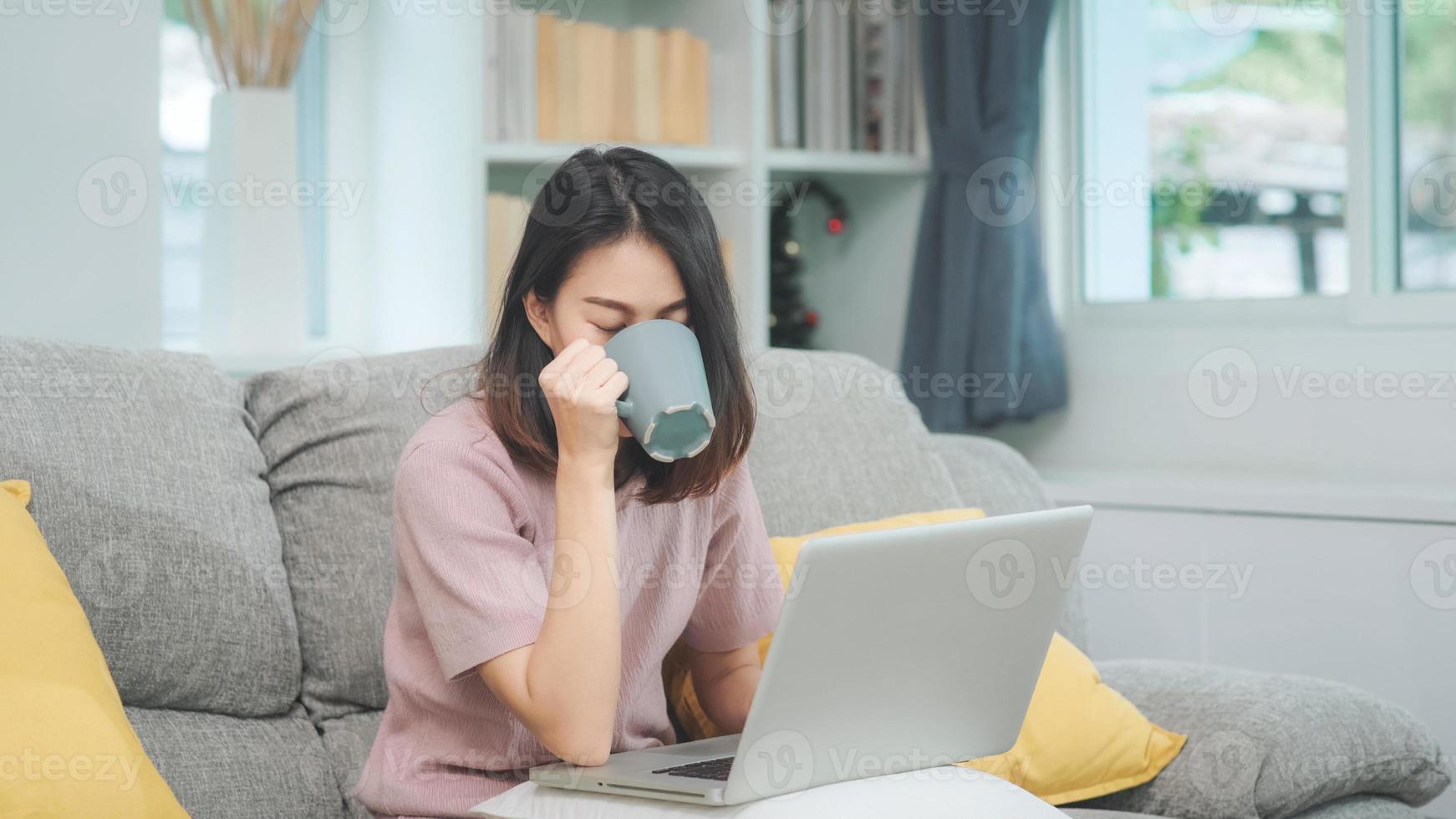 jeune femme asiatique indépendante travaillant sur un ordinateur portable vérifiant les médias sociaux et buvant du café en position allongée sur le canapé pour se détendre dans le salon à la maison. femmes de style de vie au concept de maison. photo