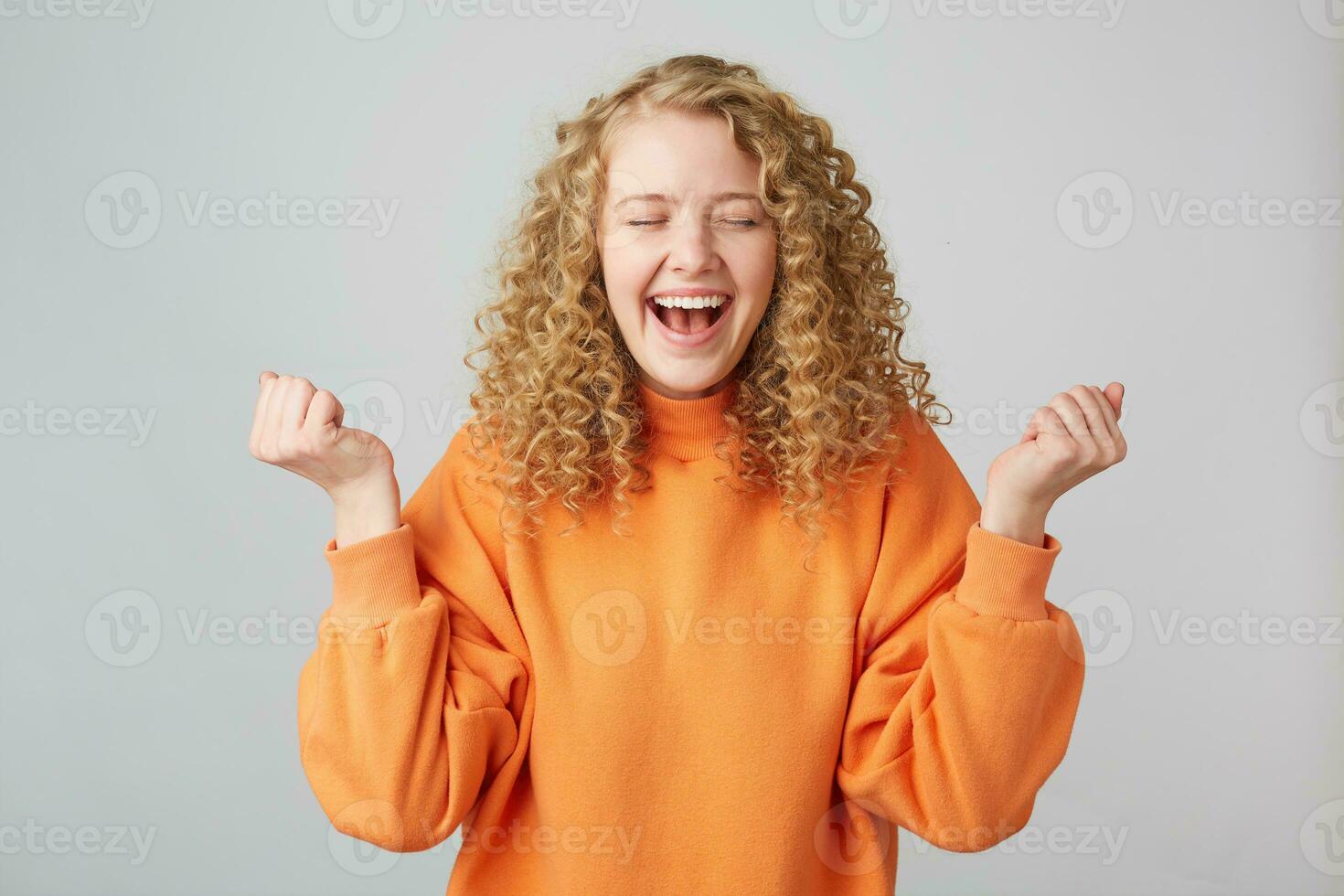 horizontal portrait de une de bonne humeur content blond femme dans Orange chandail souriant et serrant les poings comme gagnant avec yeux fermé dans plaisir, isolé plus de blanc Contexte photo