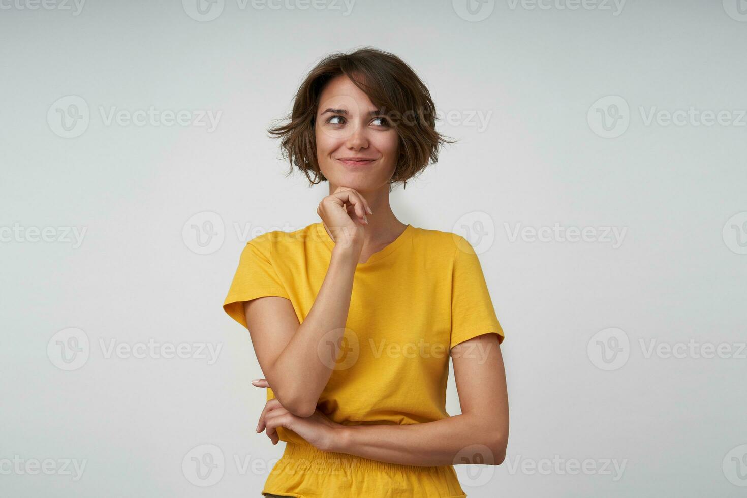 heureux jolie Jeune brunette femelle avec décontractée coiffure penché sa menton sur élevé main et à la recherche de côté positivement, posant plus de blanc Contexte dans Jaune T-shirt photo