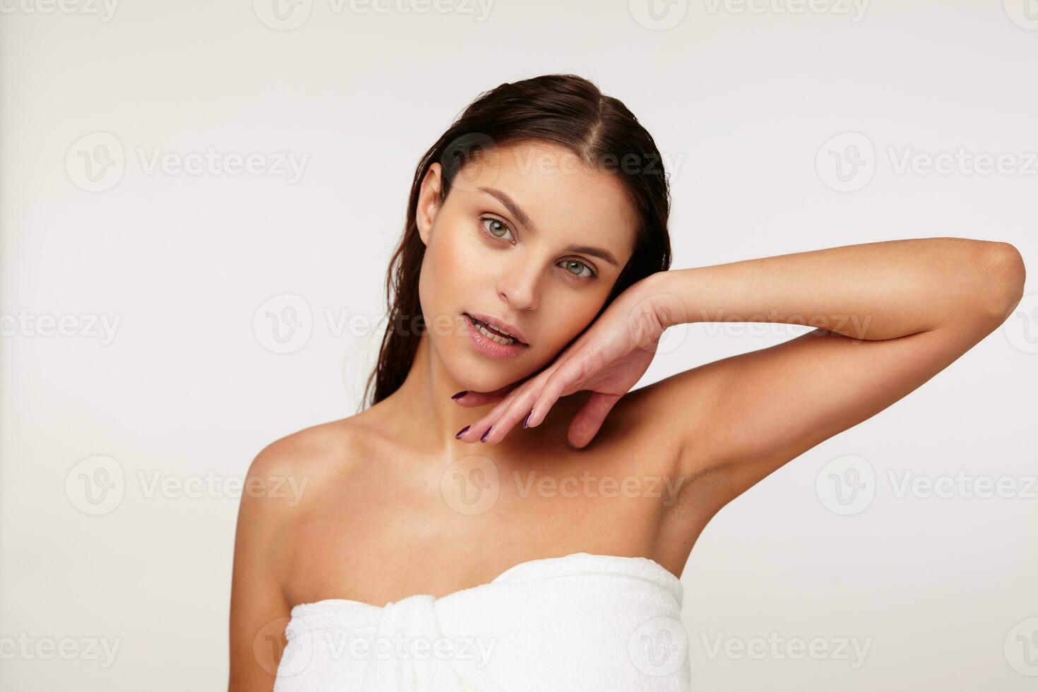 attrayant Jeune brunette aux yeux verts femelle avec humide cheveux émouvant doucement sa visage avec élevé main et à la recherche affectueusement à caméra, posant plus de blanc Contexte dans une baignoire serviette photo