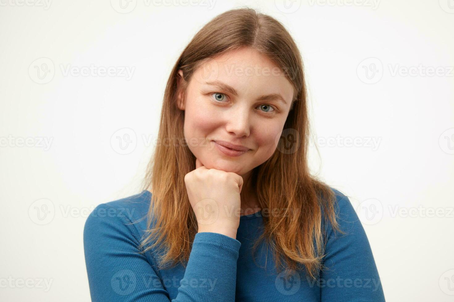 heureux Jeune femelle avec ample cheveux penché menton sur élevé main et souriant bien à caméra tandis que posant plus de blanc Contexte dans décontractée bleu chemisier photo