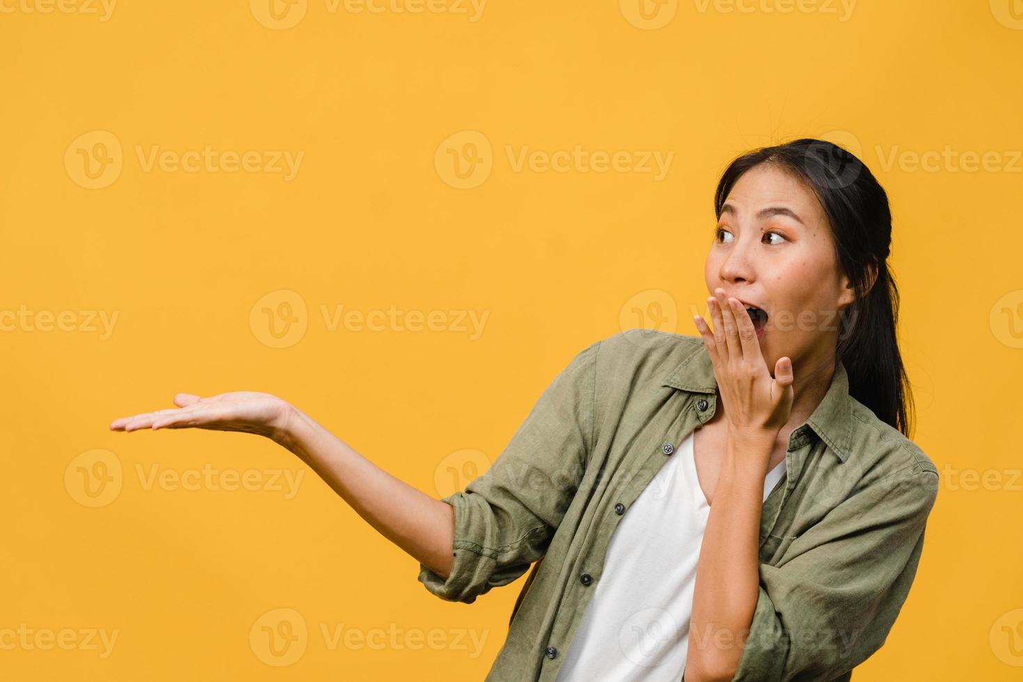 portrait d'une jeune femme asiatique souriante avec une expression joyeuse, montre quelque chose d'étonnant dans un espace vide dans des vêtements décontractés et debout isolé sur fond jaune. concept d'expression faciale. photo