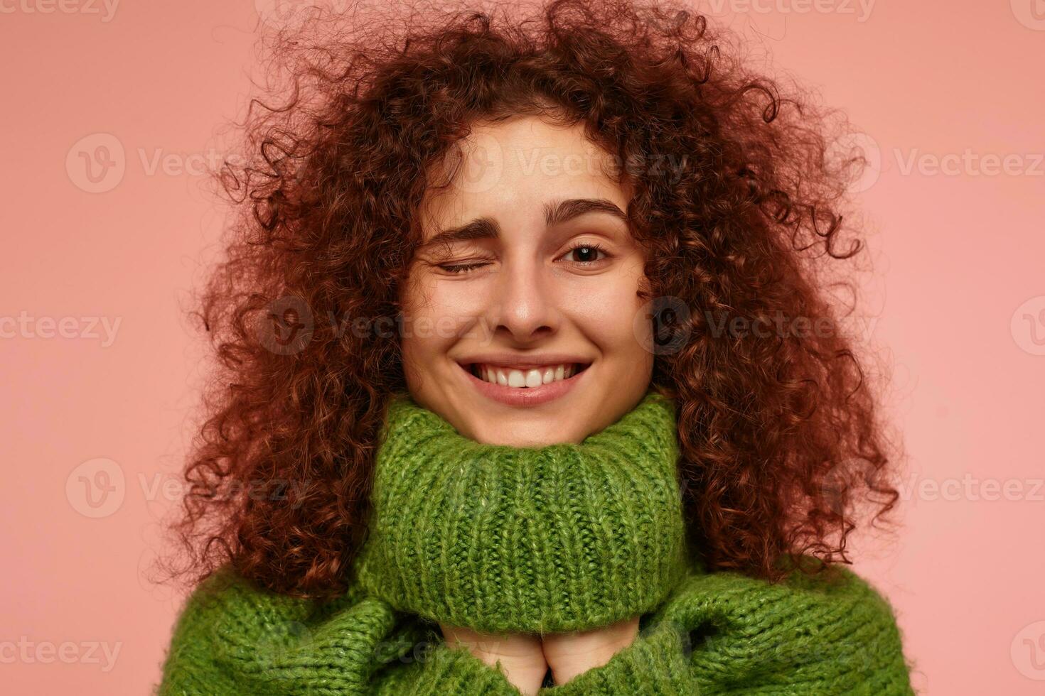 Jeune dame, jolie femme avec gingembre frisé cheveux. portant vert col roulé chandail et mettre mains en dessous de il. en train de regarder à le caméra et clin d'œil, isolé, fermer plus de pastel rose Contexte photo