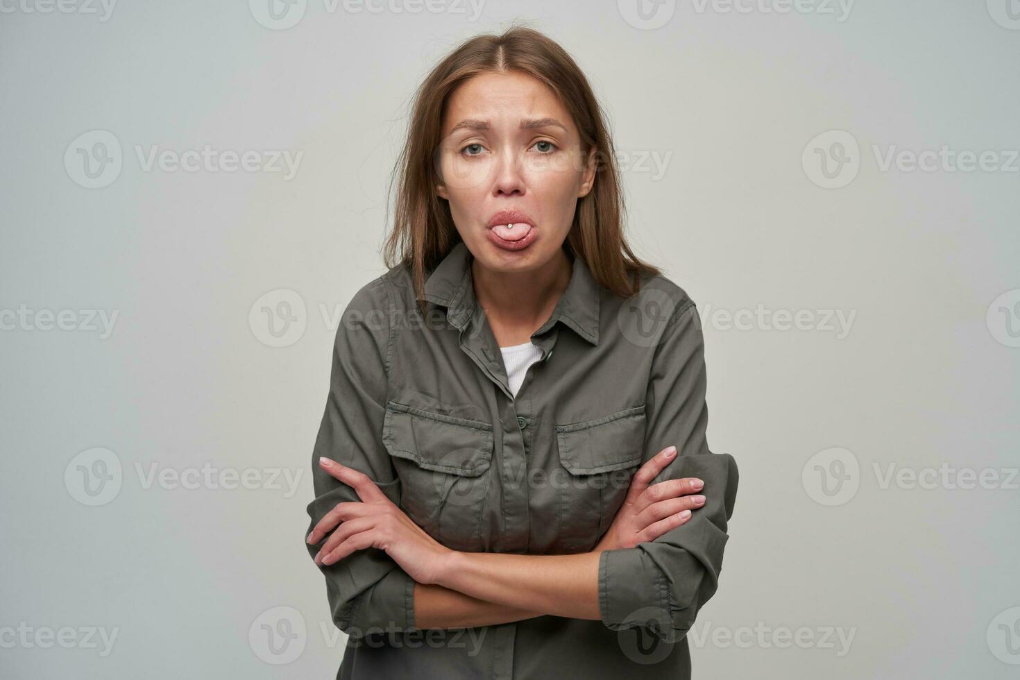 adolescent fille, dérangé à la recherche femme avec marron longue cheveux et perçant. portant gris chemise et détient bras franchi sur une poitrine. en train de regarder à le caméra et spectacles langue, isolé plus de gris Contexte photo