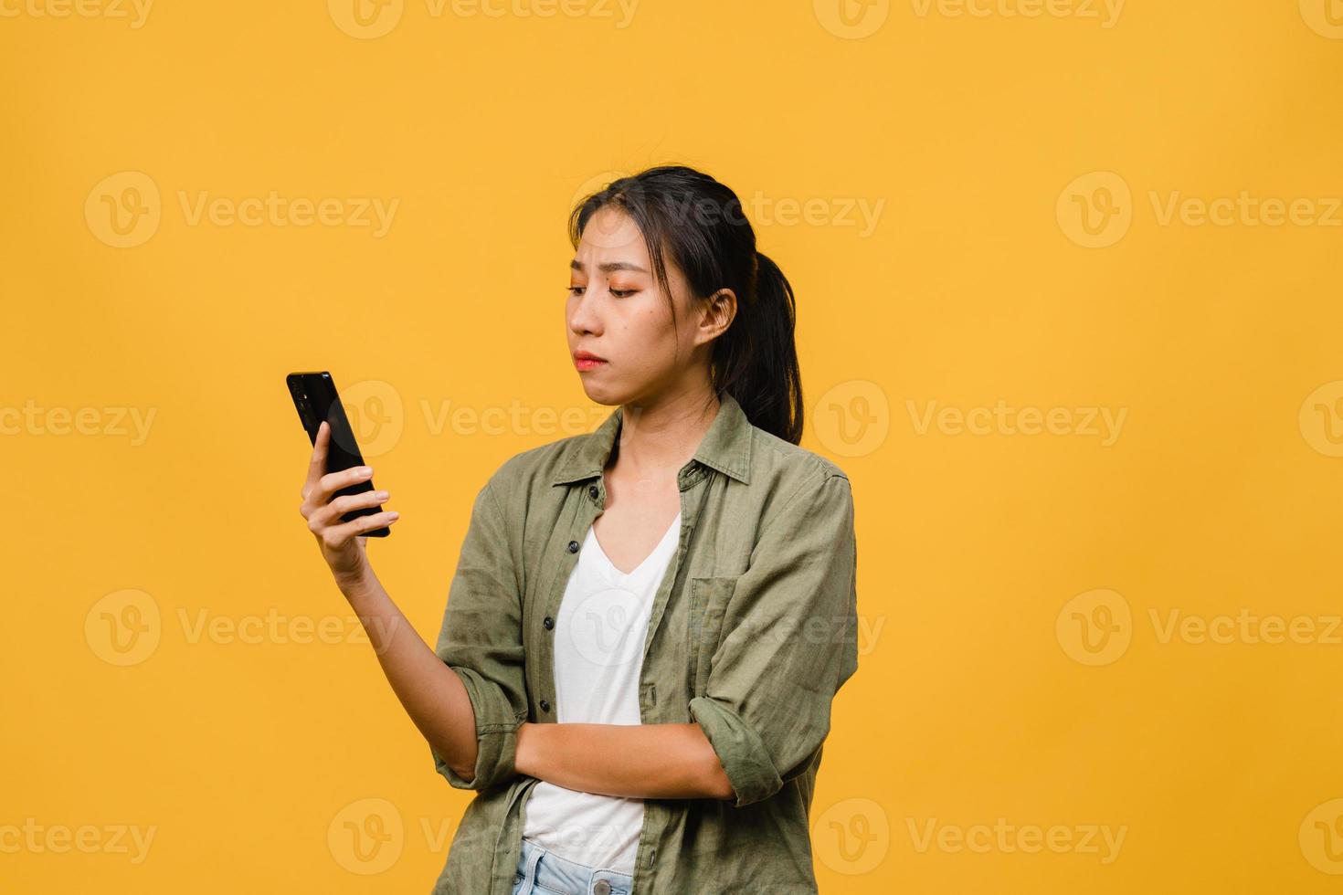 pensant rêver d'une jeune femme asiatique utilisant un téléphone avec une expression positive, vêtue d'un tissu décontracté, se sentant heureuse et se tenant isolée sur fond jaune. heureuse adorable femme heureuse se réjouit du succès. photo