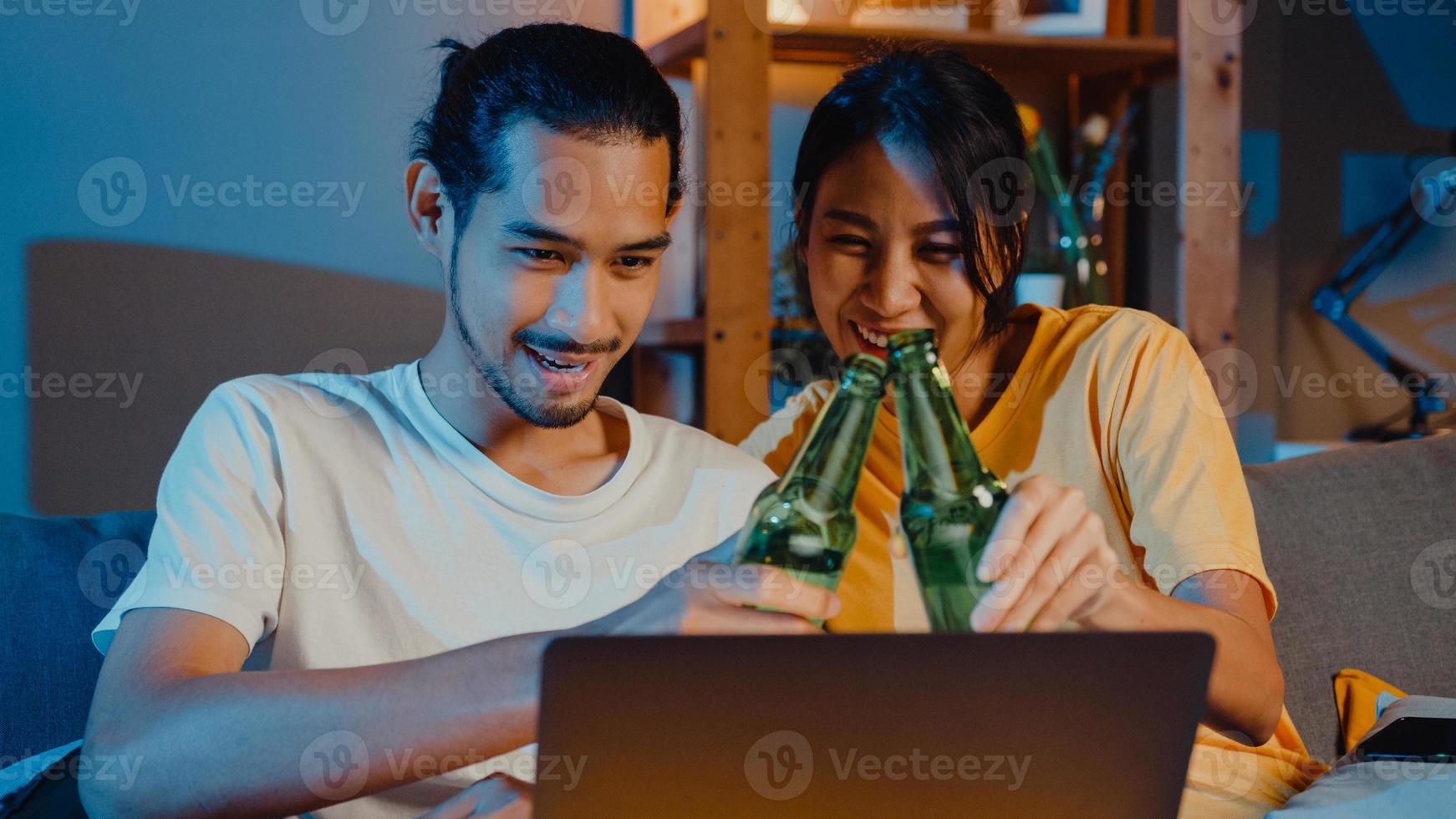 heureux jeune couple asiatique profiter d'un événement de soirée en ligne assis sur un canapé utiliser un ordinateur portable appel vidéo avec des amis boire de la bière via un appel vidéo en ligne dans le salon à la maison, concept de distanciation sociale. photo