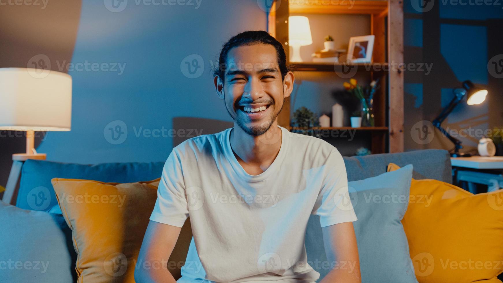 heureux jeune homme asiatique indépendant regardant la caméra sourire et parler avec des amis lors d'un appel vidéo en ligne la nuit dans le salon à la maison, rester à la maison en quarantaine, travailler à domicile, concept de distanciation sociale. photo