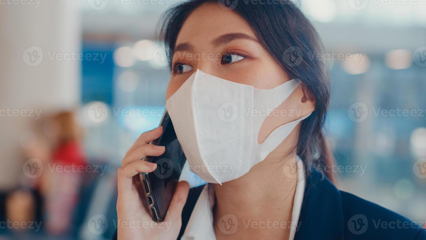 Une fille d'affaires asiatique intelligente porte un costume assis avec une valise sur un banc, appelez un téléphone intelligent avec un partenaire, attendez le vol à l'aéroport. navetteur de voyage d'affaires dans la pandémie de covid, concept de voyage d'affaires. photo