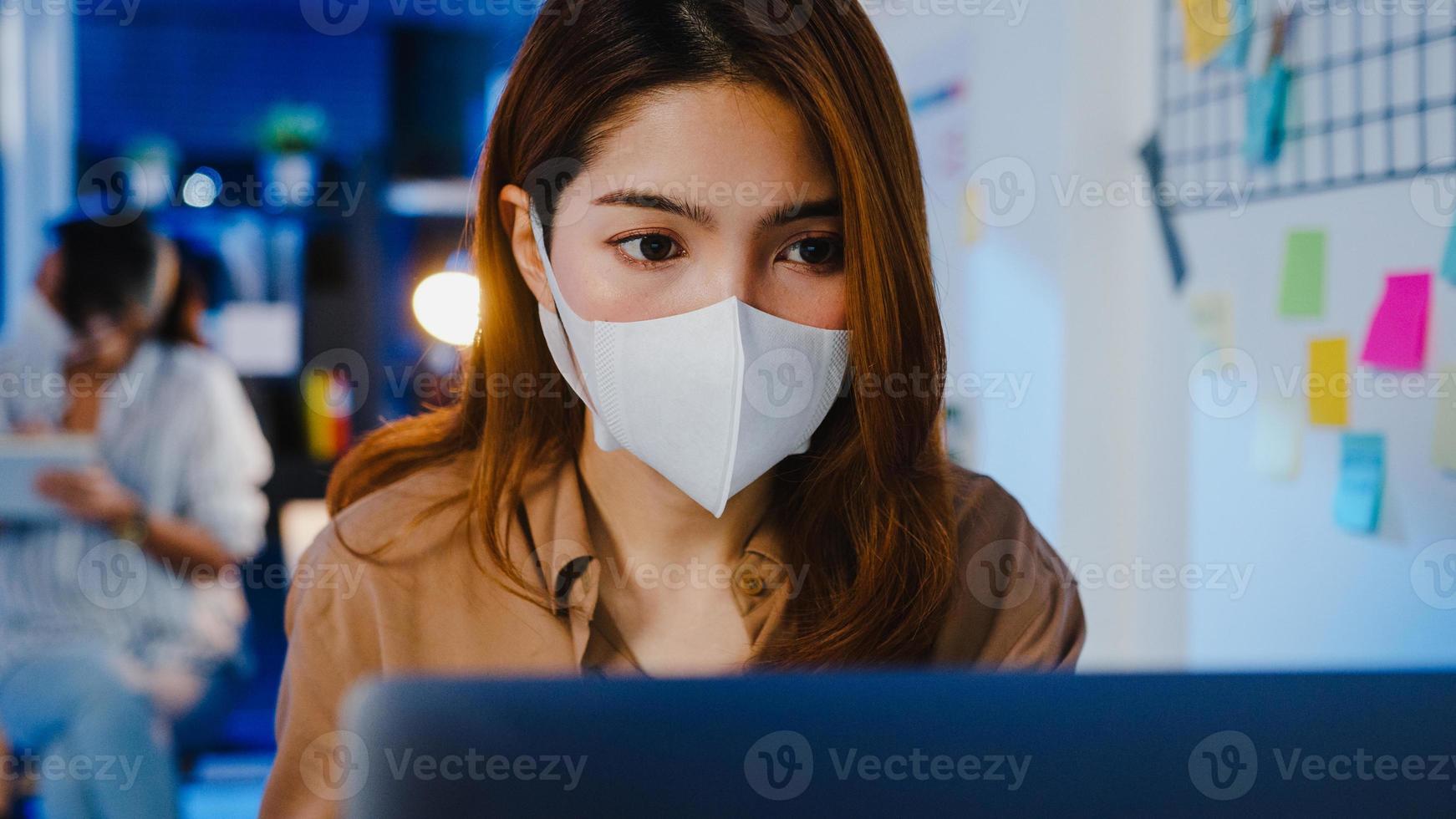 femme d'affaires asiatique heureuse portant un masque médical pour la distanciation sociale dans une nouvelle situation normale pour la prévention des virus tout en utilisant un ordinateur portable au travail la nuit au bureau. vie et travail après le coronavirus. photo