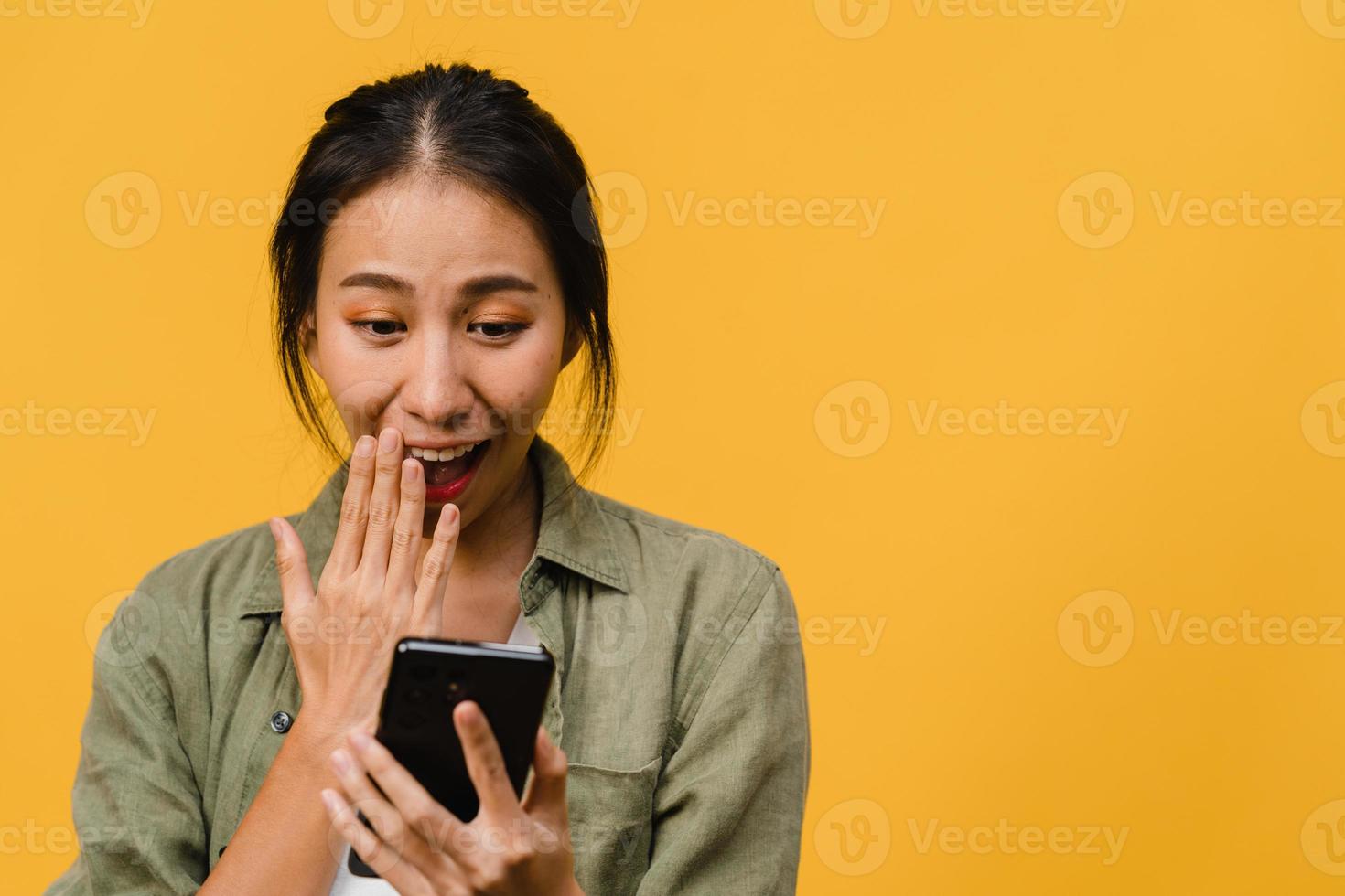 surpris une jeune femme asiatique utilisant un téléphone portable avec une expression positive, sourit largement, vêtue de vêtements décontractés et se tient isolée sur fond jaune. heureuse adorable femme heureuse se réjouit du succès. photo