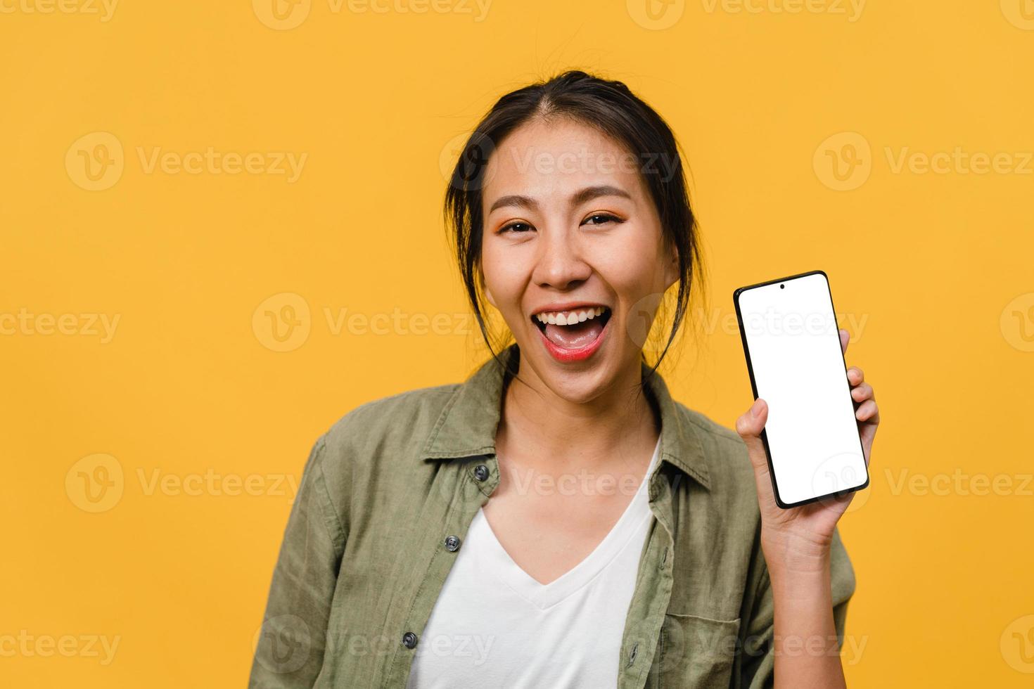 une jeune femme asiatique montre un écran de smartphone vide avec une expression positive, sourit largement, vêtue de vêtements décontractés, se sentant heureuse sur fond jaune. téléphone portable avec écran blanc en main féminine. photo