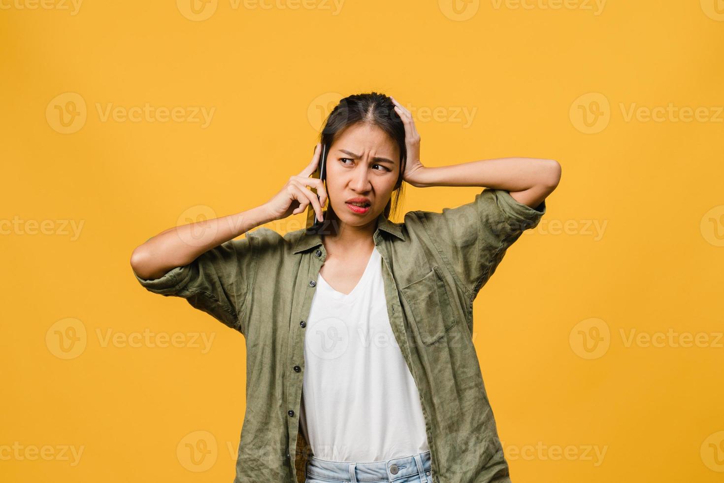 une jeune femme asiatique parle par téléphone avec une expression négative, des cris excités, des cris émotionnels en colère dans un tissu décontracté et se tient isolée sur fond jaune avec un espace de copie vierge. concept d'expression faciale. photo