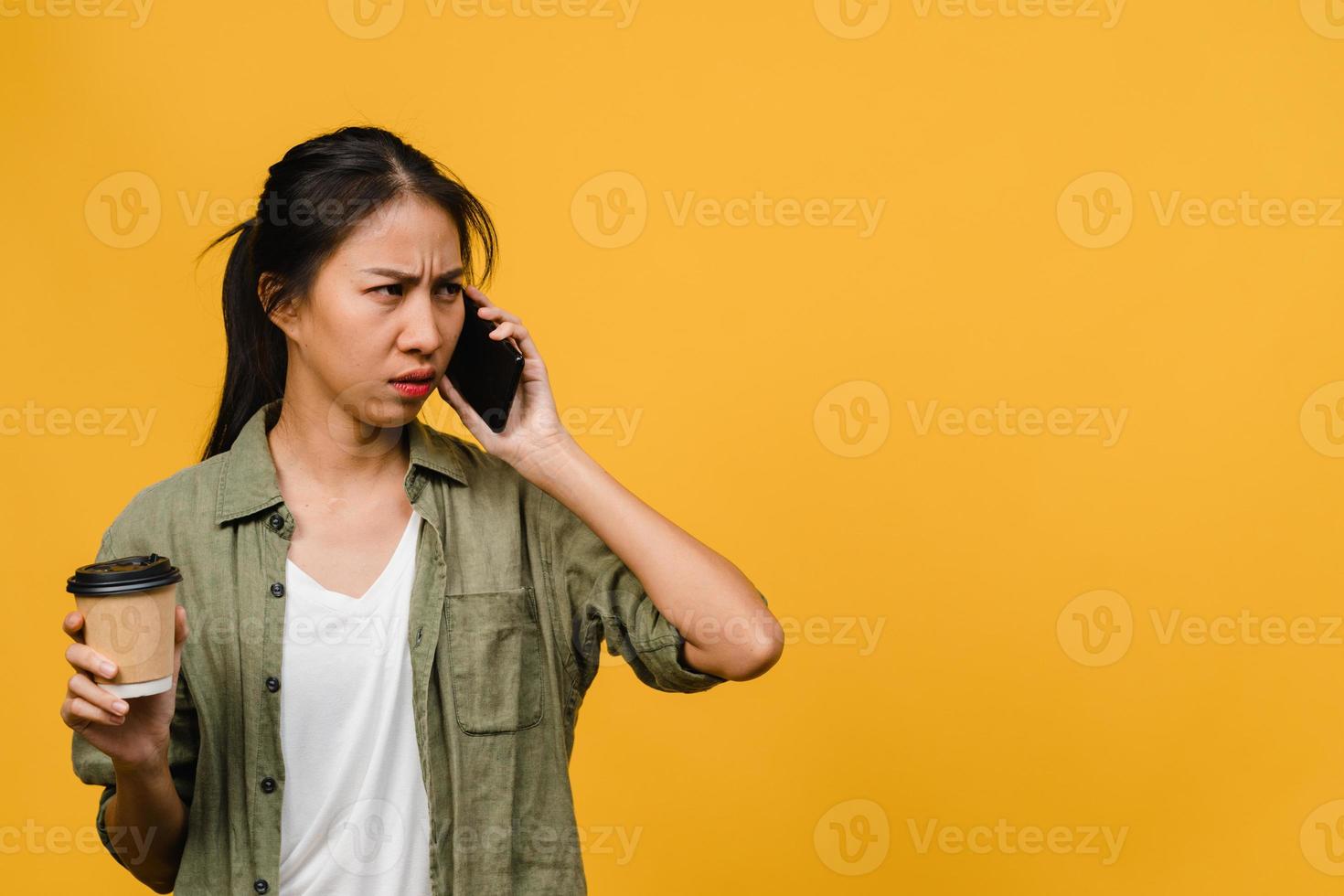 une jeune femme asiatique parle par téléphone et tient une tasse de café avec une expression négative, des cris excités, pleure émotionnellement en colère dans un tissu décontracté et se tient isolée sur fond jaune. concept d'expression faciale. photo