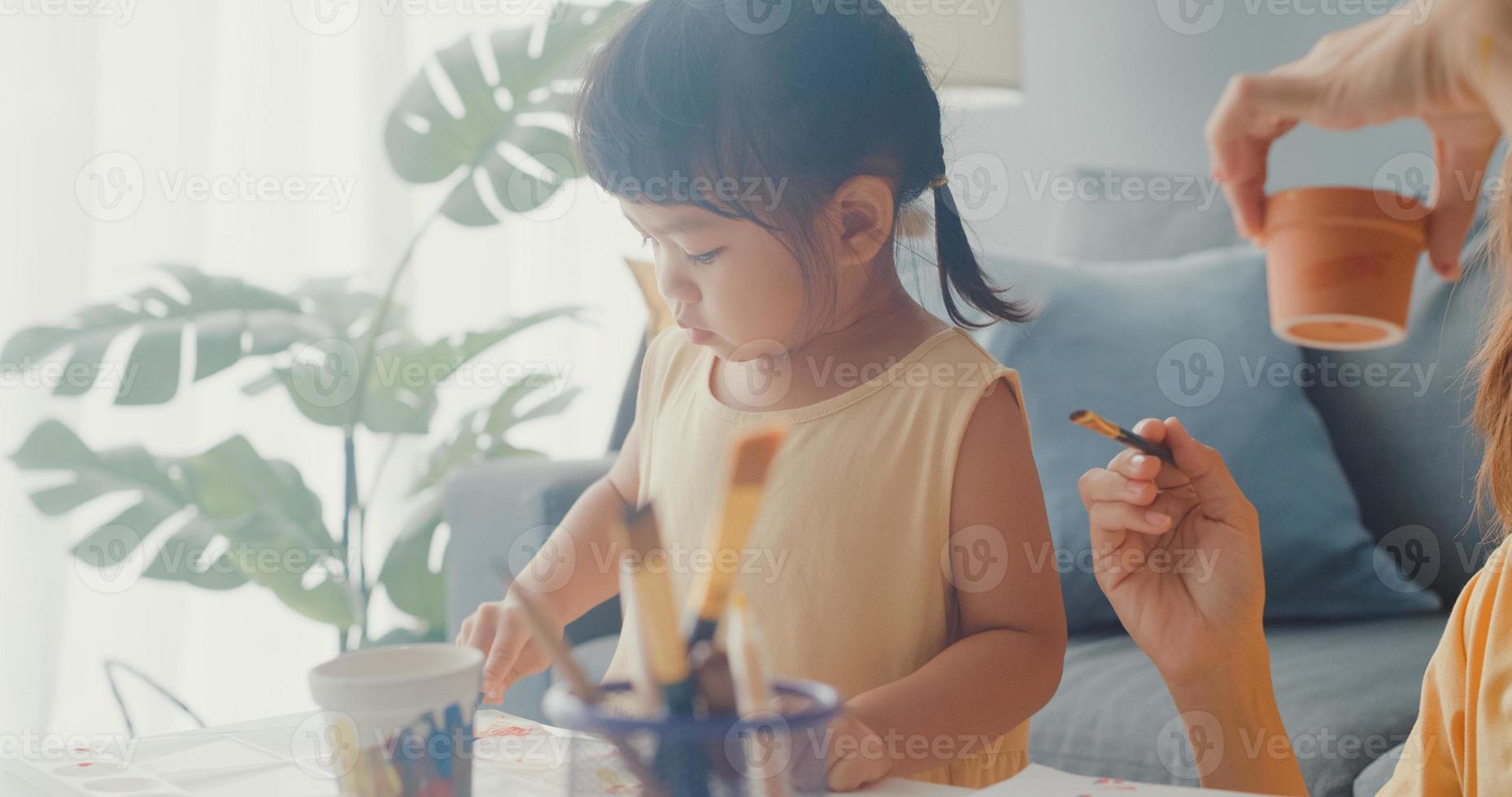 Joyeuse famille asiatique joyeuse, maman enseigne à une fille en bas âge peindre un pot en céramique s'amusant à se détendre sur une table dans le salon de la maison. passer du temps ensemble, distance sociale, quarantaine pour la prévention des coronavirus. photo