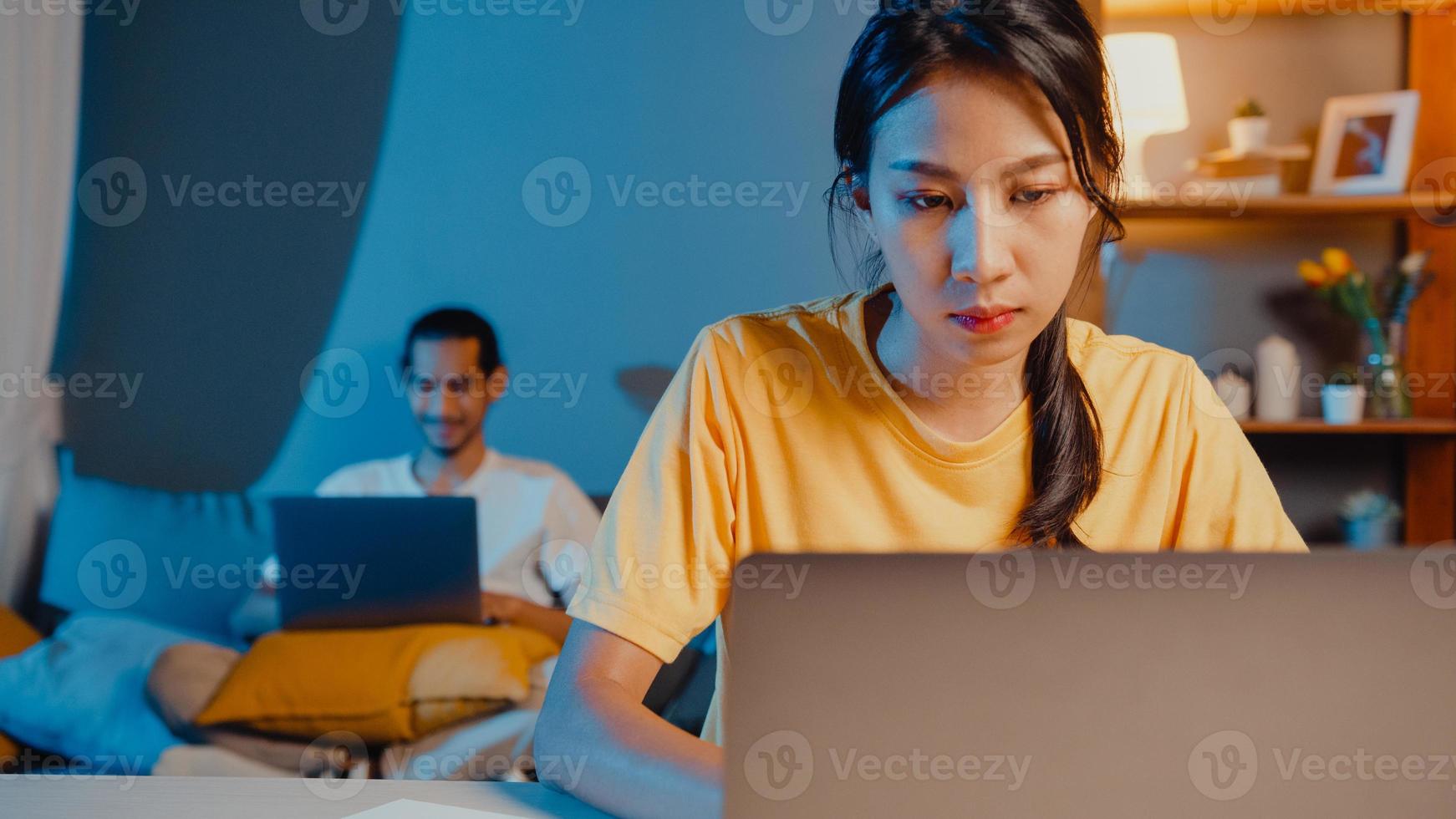 couple asiatique indépendant homme et femme dans une femme décontractée se concentre sur le travail financier dans un ordinateur portable sur le bureau la nuit un mari heureux appel vidéo avec le repos de la famille allongé après le travail sur le canapé à la maison. photo