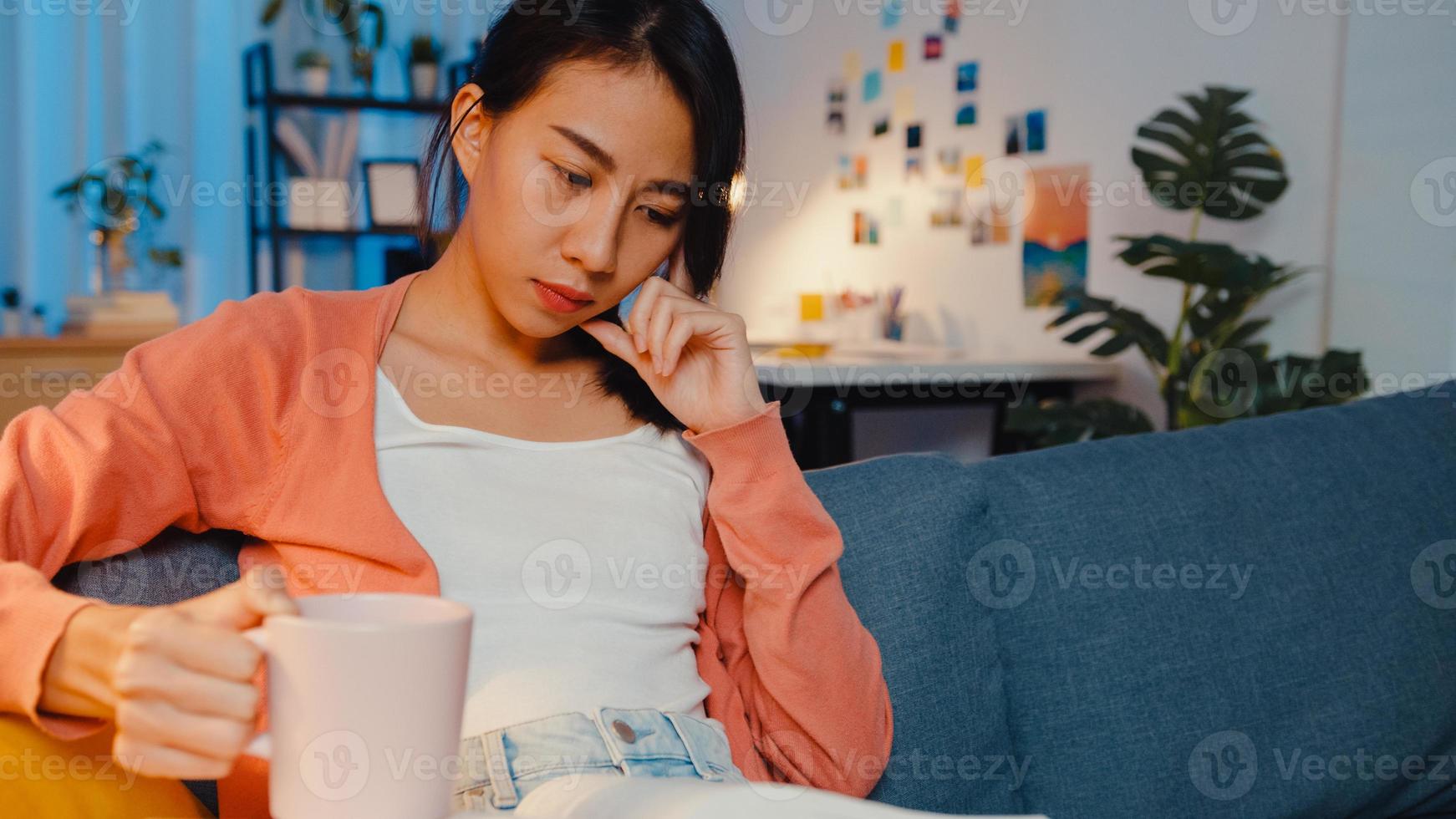 la nuit, une belle dame asiatique boit du café en se concentrant sur le livre sur le canapé du salon. éducation à domicile, rester à la maison, activité d'auto-quarantaine, activité amusante pour la quarantaine covid ou coronavirus. photo