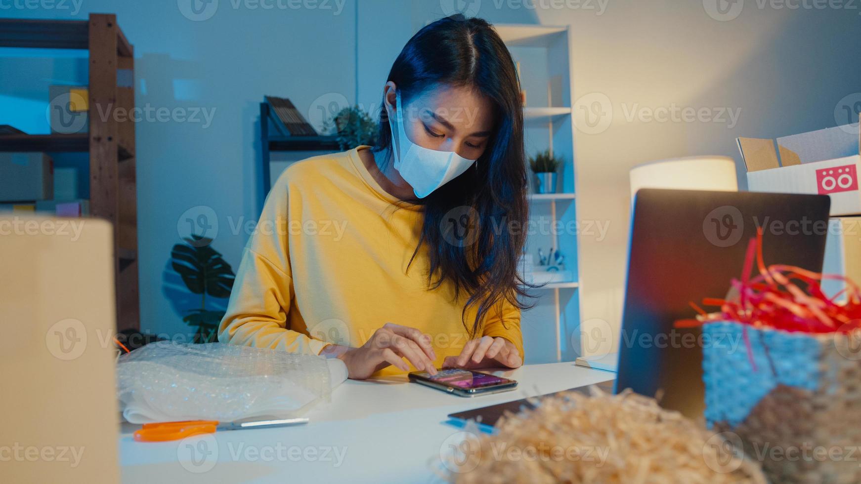 jeune femme d'affaires asiatique porter un masque facial vérifier le bon de commande sur smartphone et stocker dans l'ordinateur au bureau à domicile la nuit. propriétaire de petite entreprise, livraison sur le marché en ligne, concept de style de vie indépendant. photo