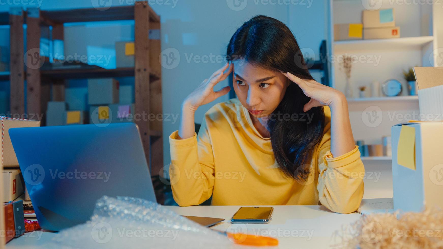 une jeune femme d'affaires asiatique regarde autour d'une pièce pleine de produits et d'une boîte à colis se sent stressée et contrariée par une mauvaise vente au bureau à domicile la nuit. propriétaire de petite entreprise, concept de livraison de marché en ligne. photo