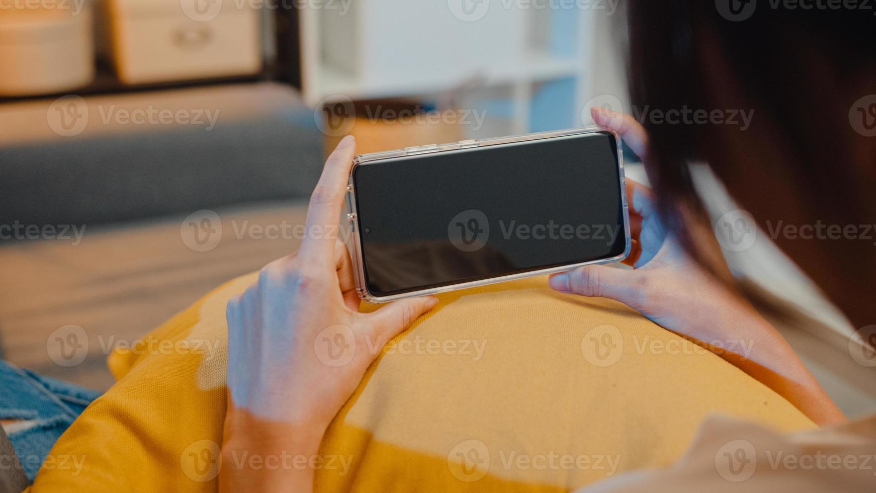 une jeune femme asiatique utilise un téléphone intelligent avec un écran noir vierge pour un texte publicitaire tout en se reposant sur un canapé dans le salon lors d'une nuit à la maison moderne. technologie de clé de chrominance, concept de design marketing. photo