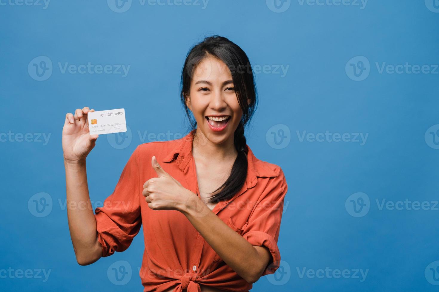 jeune femme asiatique montre une carte bancaire de crédit avec une expression positive, sourit largement, vêtue de vêtements décontractés, se sentant heureuse et isolée sur fond bleu. concept d'expression faciale. photo