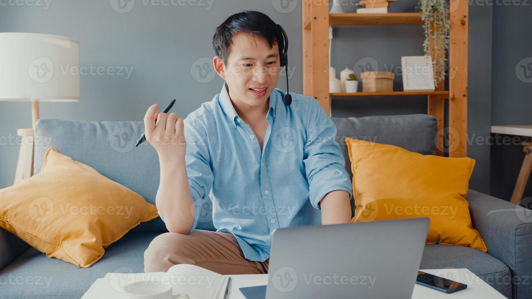 un jeune homme d'affaires asiatique porte des écouteurs à l'aide d'un ordinateur portable et parle à ses collègues du plan lors d'un appel vidéo pendant qu'il travaille à domicile dans le salon. auto-isolement, distanciation sociale, quarantaine pour la prévention du covid. photo