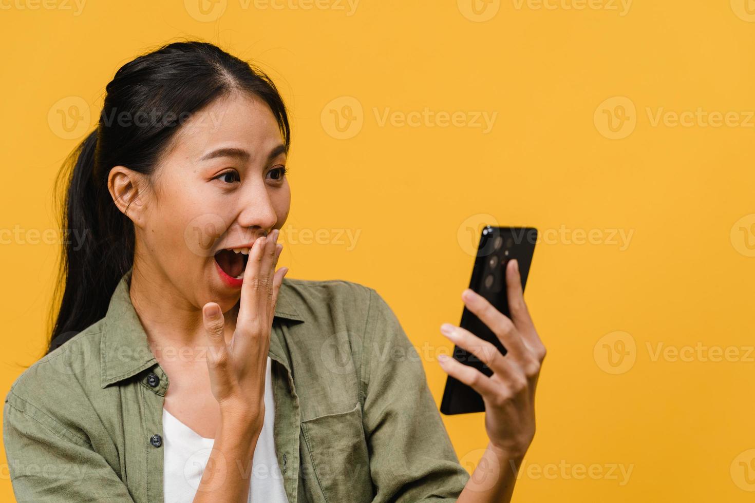 surpris une jeune femme asiatique utilisant un téléphone portable avec une expression positive, sourit largement, vêtue de vêtements décontractés et se tient isolée sur fond jaune. heureuse adorable femme heureuse se réjouit du succès. photo