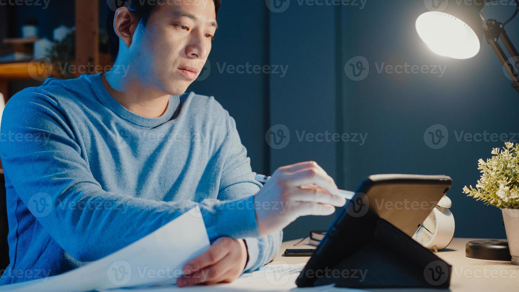 un homme d'affaires indépendant d'asie se concentre sur un stylo de travail écrit sur un ordinateur tablette occupé avec plein de documents graphiques sur le bureau dans le salon à la maison des heures supplémentaires la nuit, travail à domicile pendant le concept de pandémie de covid-19. photo