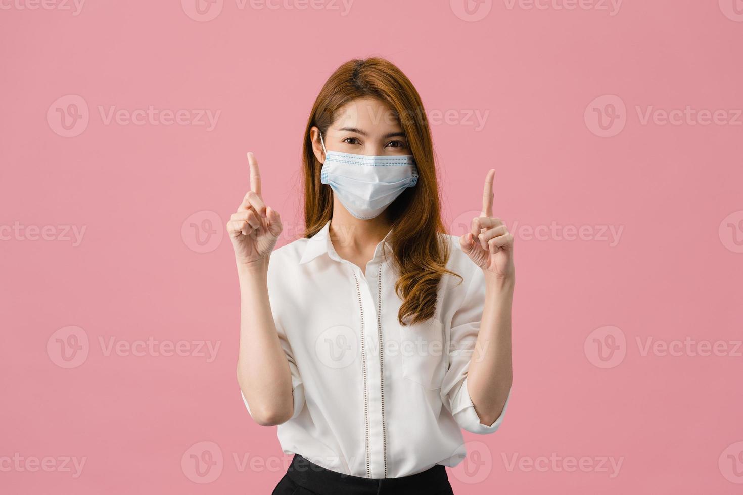 une jeune fille asiatique portant un masque médical montre quelque chose dans un espace vide vêtue d'un tissu décontracté et regardant la caméra isolée sur fond rose. distanciation sociale, quarantaine pour le virus corona. photo