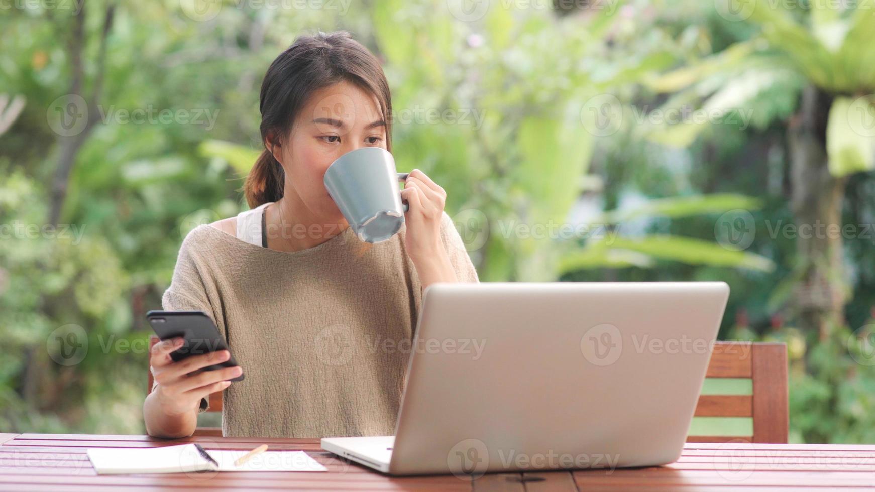 femme asiatique indépendante travaillant à la maison, femme d'affaires travaillant sur ordinateur portable et utilisant un téléphone portable buvant du café assis sur une table dans le jardin le matin. femmes de style de vie travaillant à la maison concept. photo