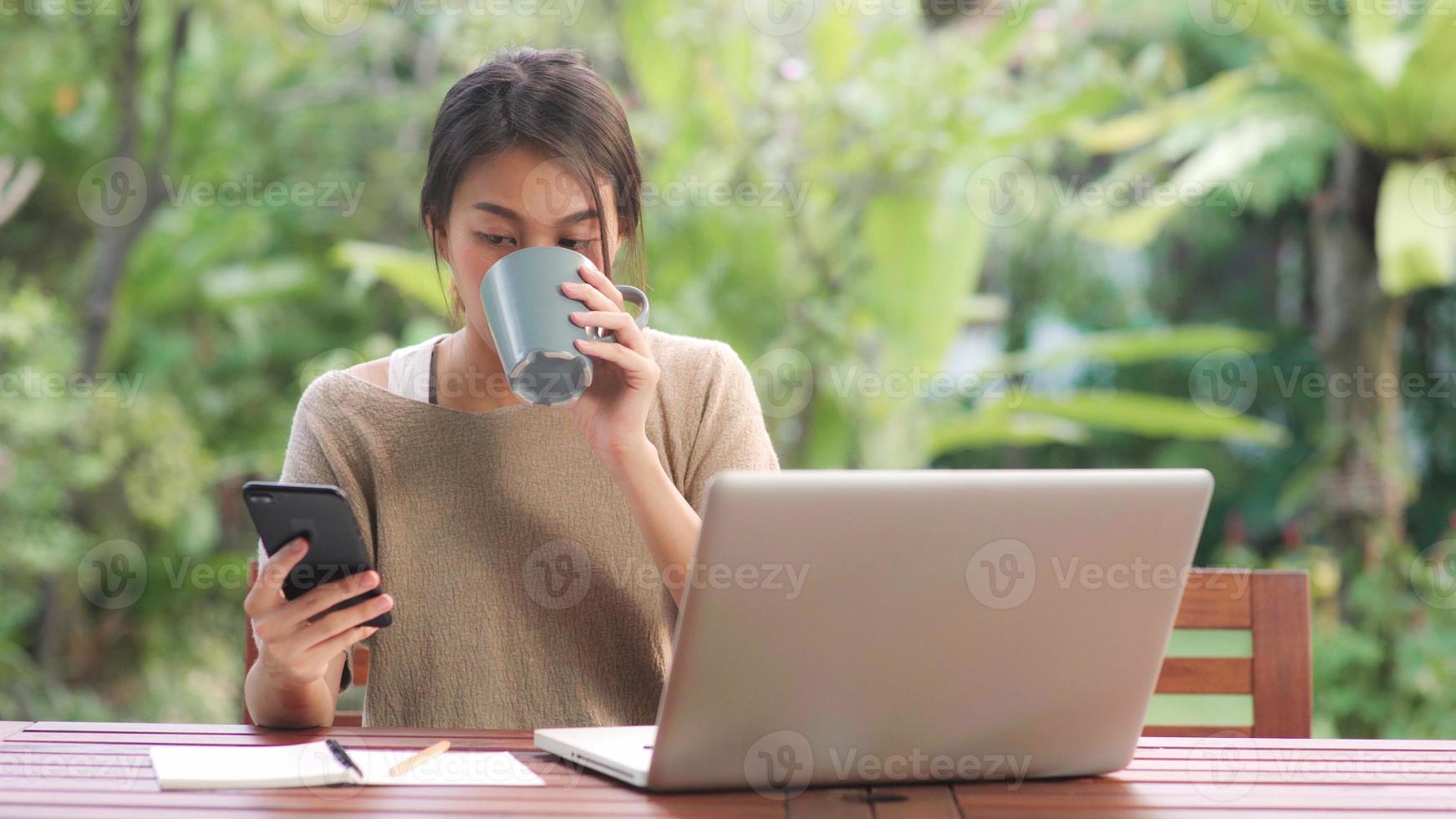 femme asiatique indépendante travaillant à la maison, femme d'affaires travaillant sur ordinateur portable et utilisant un téléphone portable buvant du café assis sur une table dans le jardin le matin. femmes de style de vie travaillant à la maison concept. photo