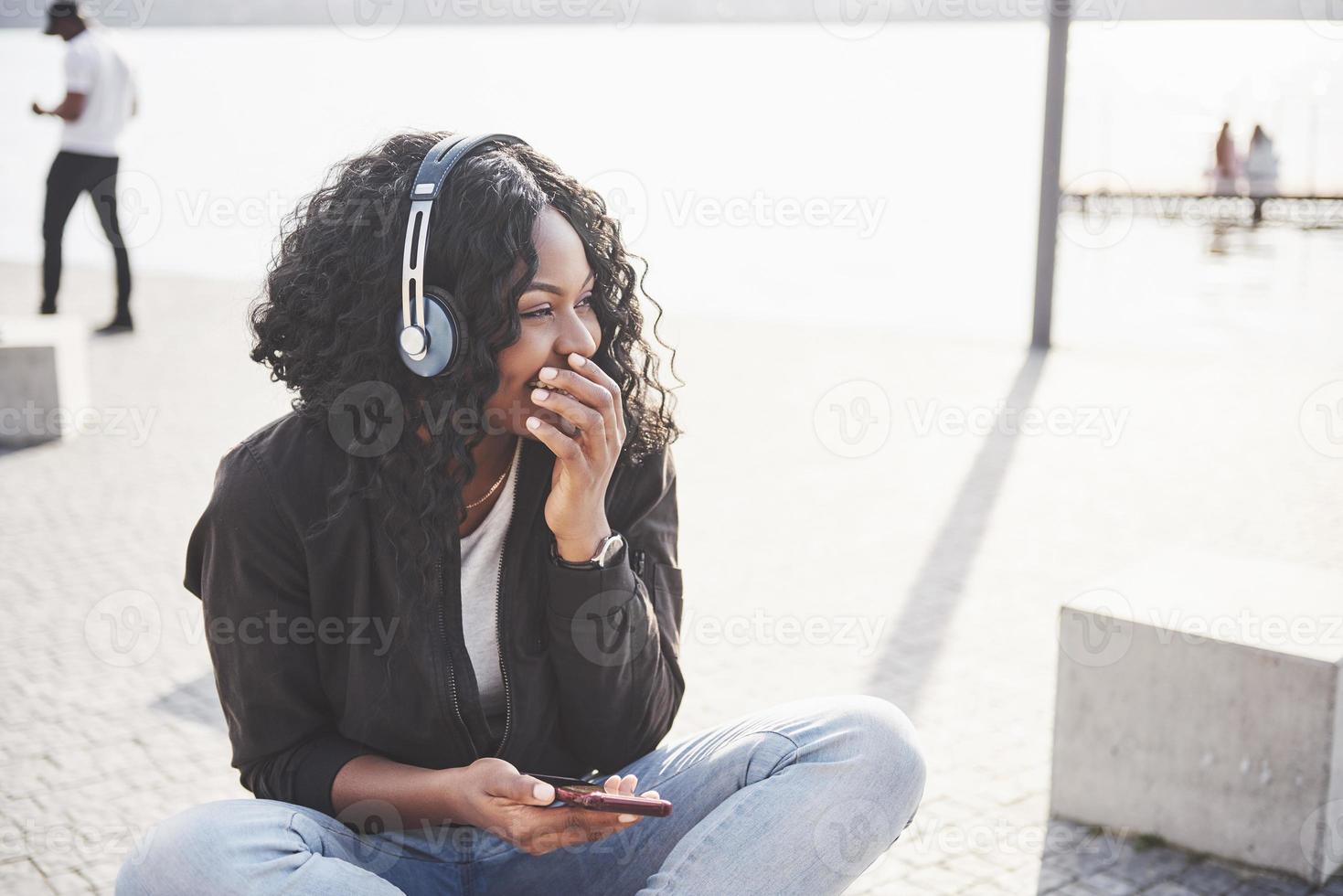 portrait d'une belle jeune jolie fille afro-américaine assise sur la plage ou le lac et écoutant de la musique dans ses écouteurs photo