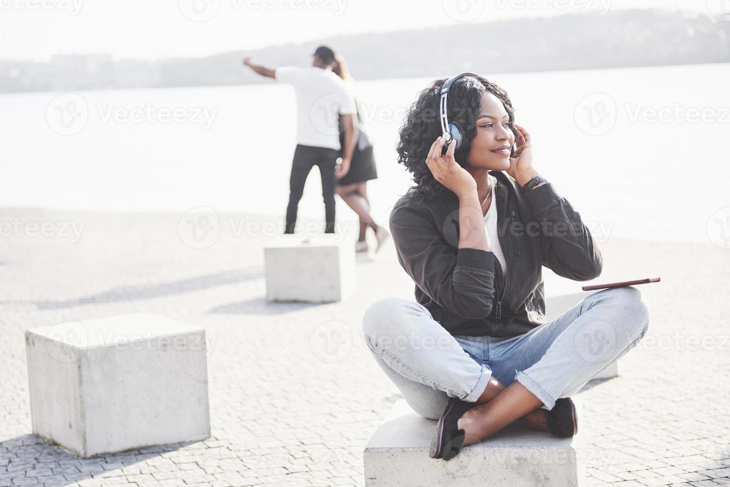 portrait d'une belle jeune jolie fille afro-américaine assise sur la plage ou le lac et écoutant de la musique dans ses écouteurs photo