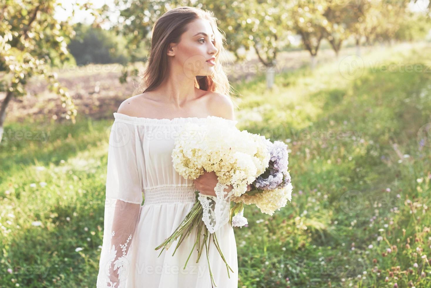 une belle jeune fille vêtue d'une robe blanche et d'un bouquet de fleurs d'été passe une belle journée dans le jardin photo