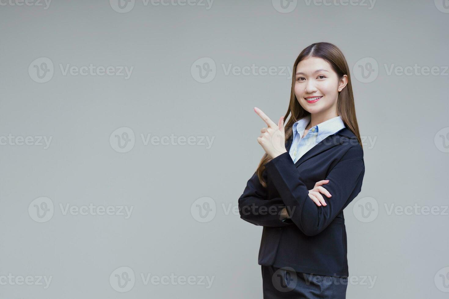 professionnel asiatique travail femme qui a longue cheveux porte noir formel costume avec bleu chemise tandis que elle bras traversée et souriant en présentant sur gris Contexte. photo