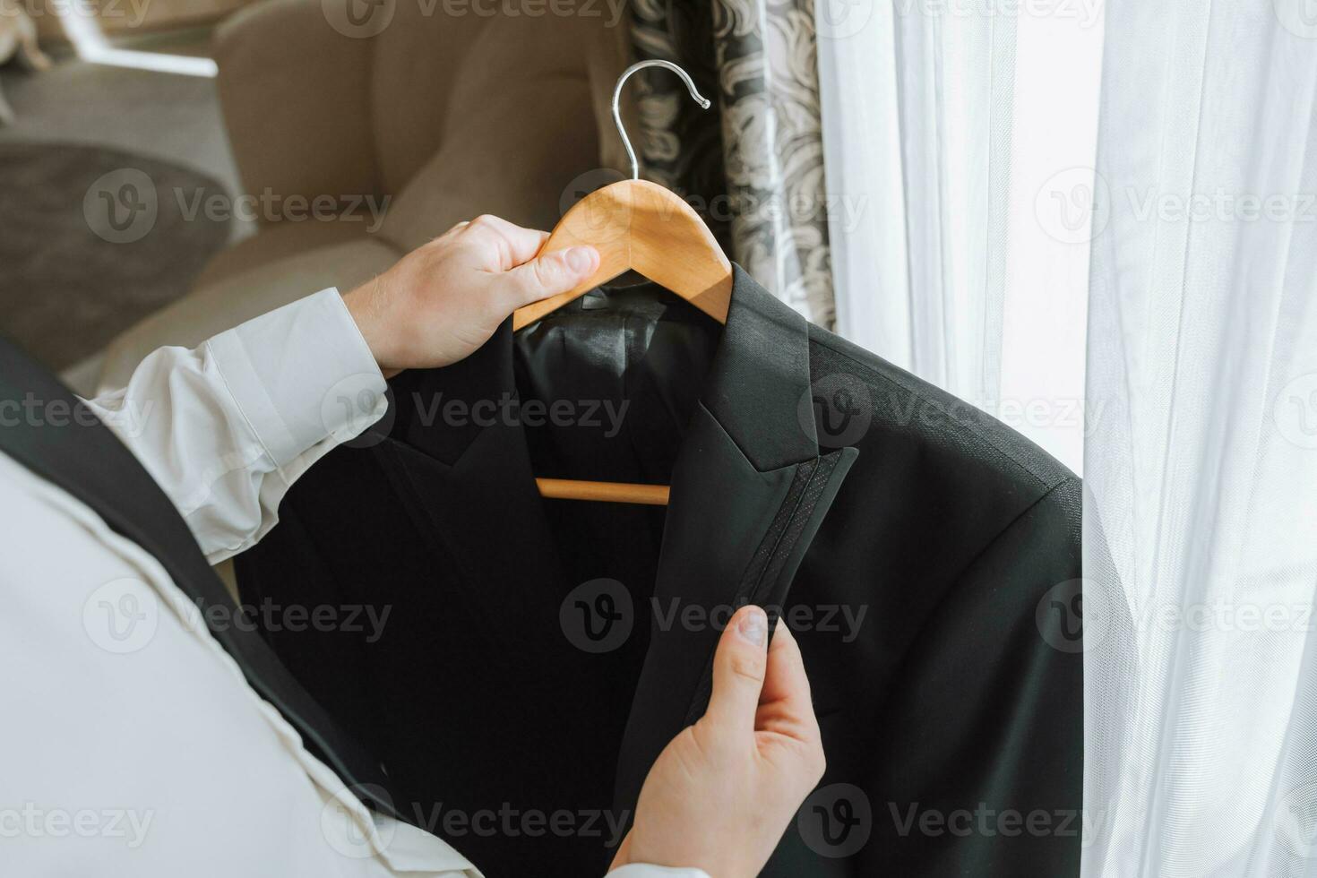 une Jeune homme détient une veste dans le sien mains dans le sien chambre, près le fenêtre dans une Hôtel chambre, fermer photo. le jeune marié est en train de préparer pour le mariage la cérémonie photo