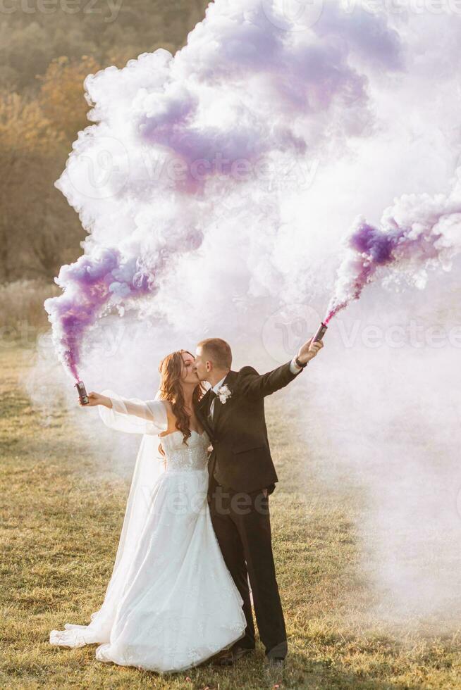 la mariée et jeune marié en jouant avec coloré fumée dans violet mains, câlins et bisous. fumée bombes à une mariage. photo