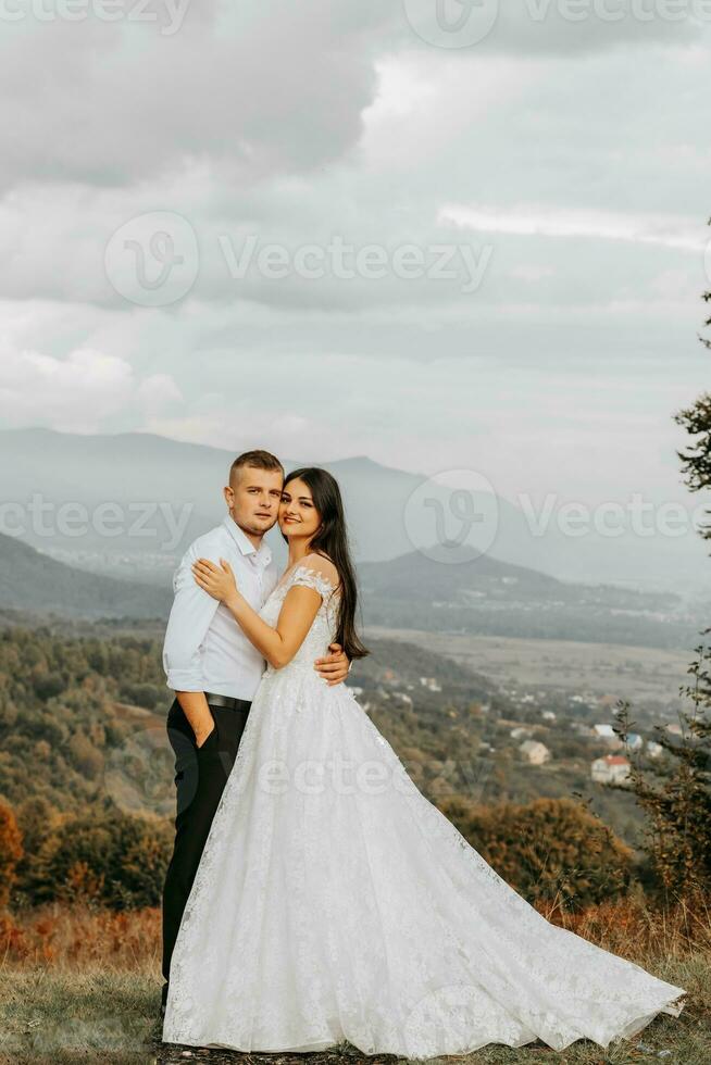 une mariage couple tendrement câlins dans le montagnes. le jeune marié câlins le la mariée par le épaules. une magnifique et magnifique de la mariée robe. photo