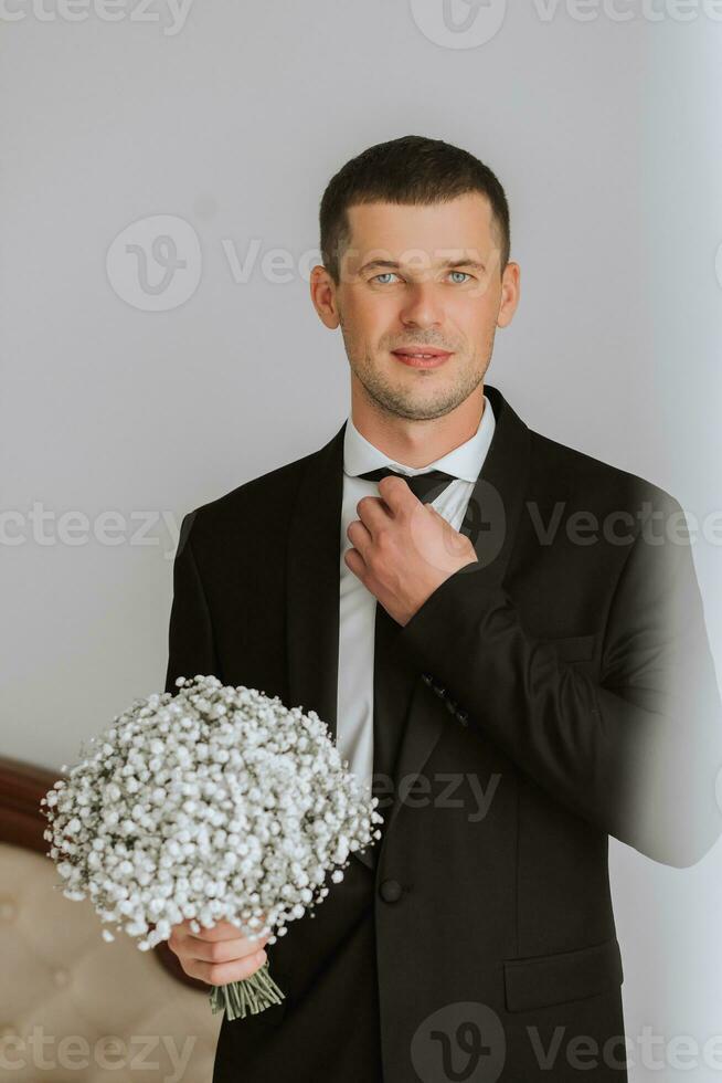 portrait de une Jeune homme dans une blanc chemise et cravate, en portant une bouquet de gypsophile fleurs. le jeune marié est en train de préparer pour le mariage cérémonie. photo