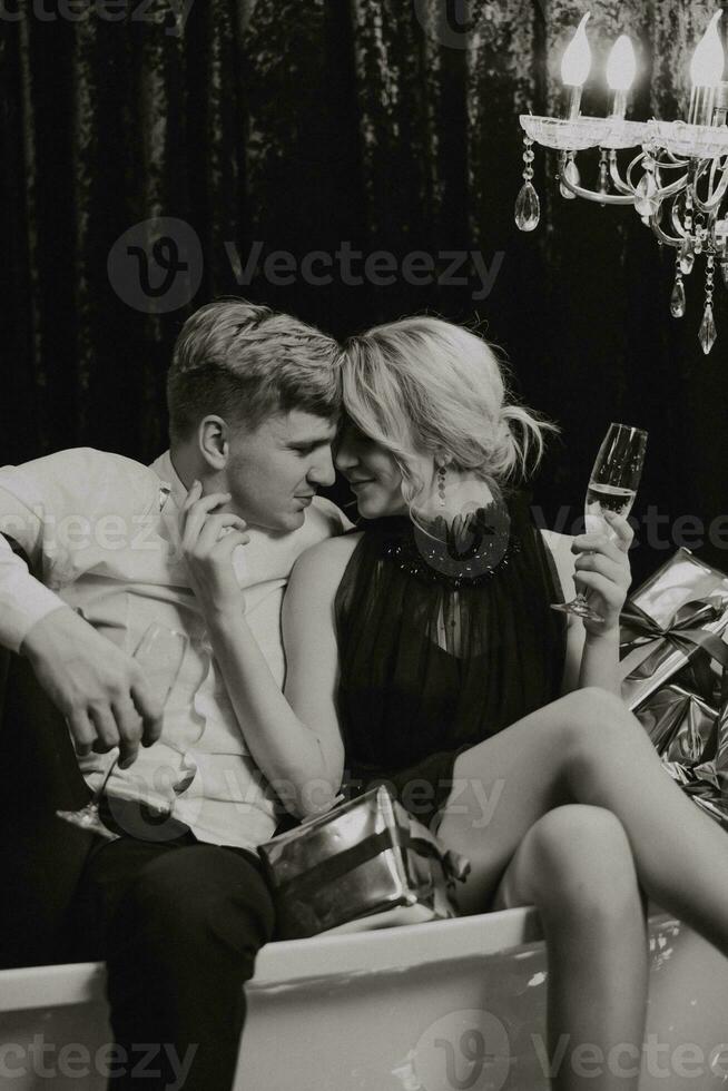 une romantique femme est mensonge dans le salle de bains avec Nouveau années cadeaux, sa mari est permanent suivant à sa avec des lunettes de Champagne. joyeux Noël. noir et blanc photo avec artistique des bruits