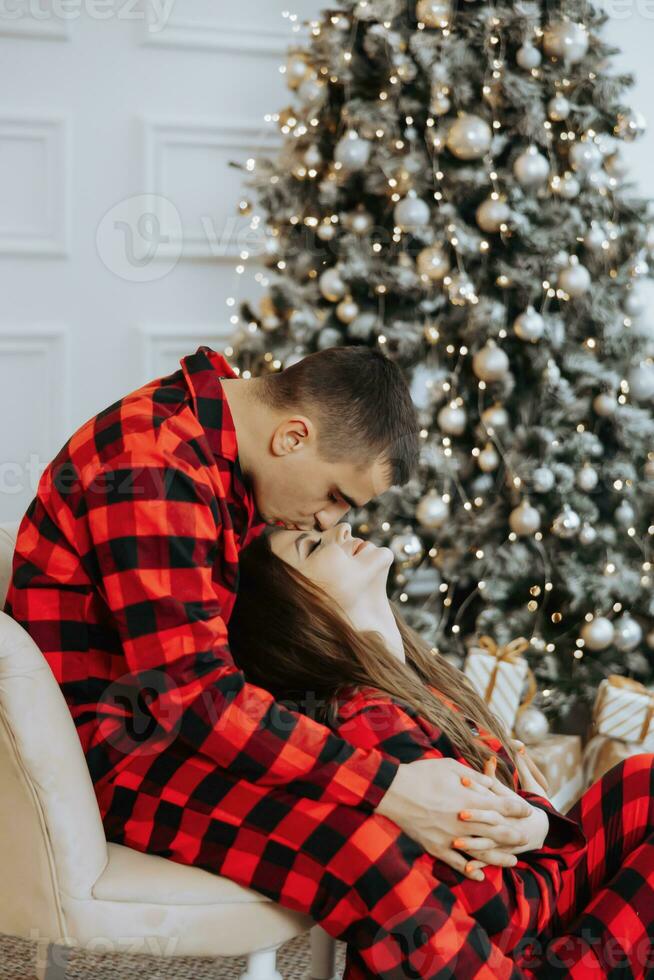 Jeune magnifique couple dans correspondant à rouge pyjamas près le Noël arbre. joie, câlins et bisous. Nouveau années vacances et cadeaux en dessous de le Noël arbre photo